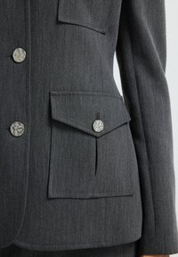 Blazer gris ajustado con una textura suave, dos bolsillos con solapas y botones metálicos con un diseño estampado.