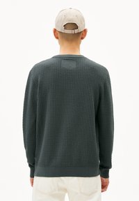 Pull vert foncé avec un design texturé en maille gaufrée, des poignets et un ourlet côtelés. Présente une petite étiquette rectangulaire à l'arrière.