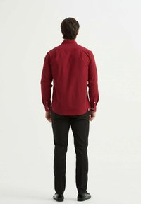 Chemise rouge à manches longues avec poignets à boutons et col pointu, associée à un pantalon noir et des chaussures, vue de dos.