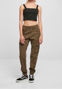 Woman wearing a black cropped tank top, brown cargo pants with side pockets, and black-and-white sneakers, standing against a plain background.