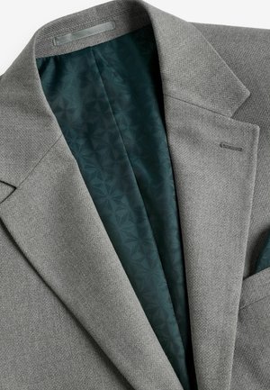 Grauer Blazer aus strukturiertem Stoff. Innenausstattung mit einem gemusterten dunkelgrünen Futter. Auffällige Details sind der geschlitzte Revers und die Tasche.