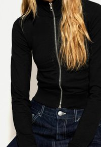 Czarna kurtka zapinana na zamek z wysokim kołnierzem, dopasowanymi długimi rękawami i prążkowanym dołem. Do tego ciemnoniebieskie dżinsy z srebrnym guzikiem.