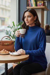 Donna con maglione blu seduta vicino a una finestra, che tiene una tazza bianca, con un cornetto su un piatto e una borsa marrone sul tavolo.