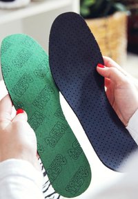 Green and black insoles with textured surfaces, featuring perforations. The green side has a printed pattern, while the black side is smooth.