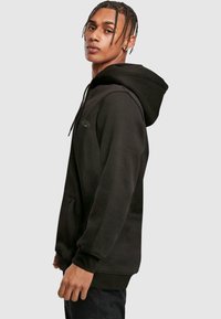 Sudadera negra con capucha, bolsillo canguro, puños acanalados y una textura suave. Presenta un sutil logo en el pecho, que resalta un ajuste relajado.