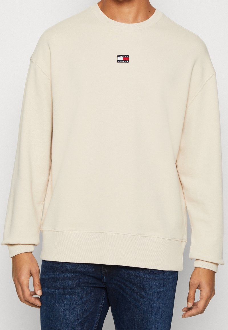 Uomo che indossa una felpa crema con collo rotondo e un piccolo logo Tommy Jeans sul petto, insieme a jeans blu scuro, in piedi davanti a uno sfondo bianco.