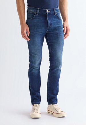 Homme debout portant un jean slim bleu foncé et des baskets en toile écrues avec lacets sur fond uni.