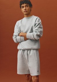 Jeune homme aux cheveux bouclés portant un sweat-shirt gris clair et un short assorti, debout les bras croisés devant un fond marron.
