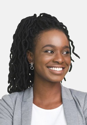 Lächelnde Frau mit schwarzen Dreadlocks, die ein hellgraues Blazer über einem weißen Oberteil und silbernen Creolen trägt. Einfarbiger weißer Hintergrund.