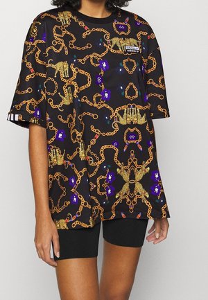 Mujer con pantalones cortos negros y una camiseta negra holgada con cadenas doradas, flores moradas y estampados de leopardo, con un logo de Adidas.