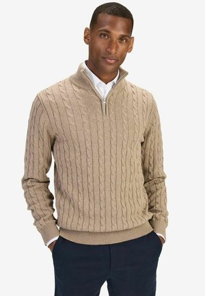 Beige kabelstickad tröja med halvzip-krage, med texturerade mönster och ribbade ärmslut, i kombination med marinblå byxor.