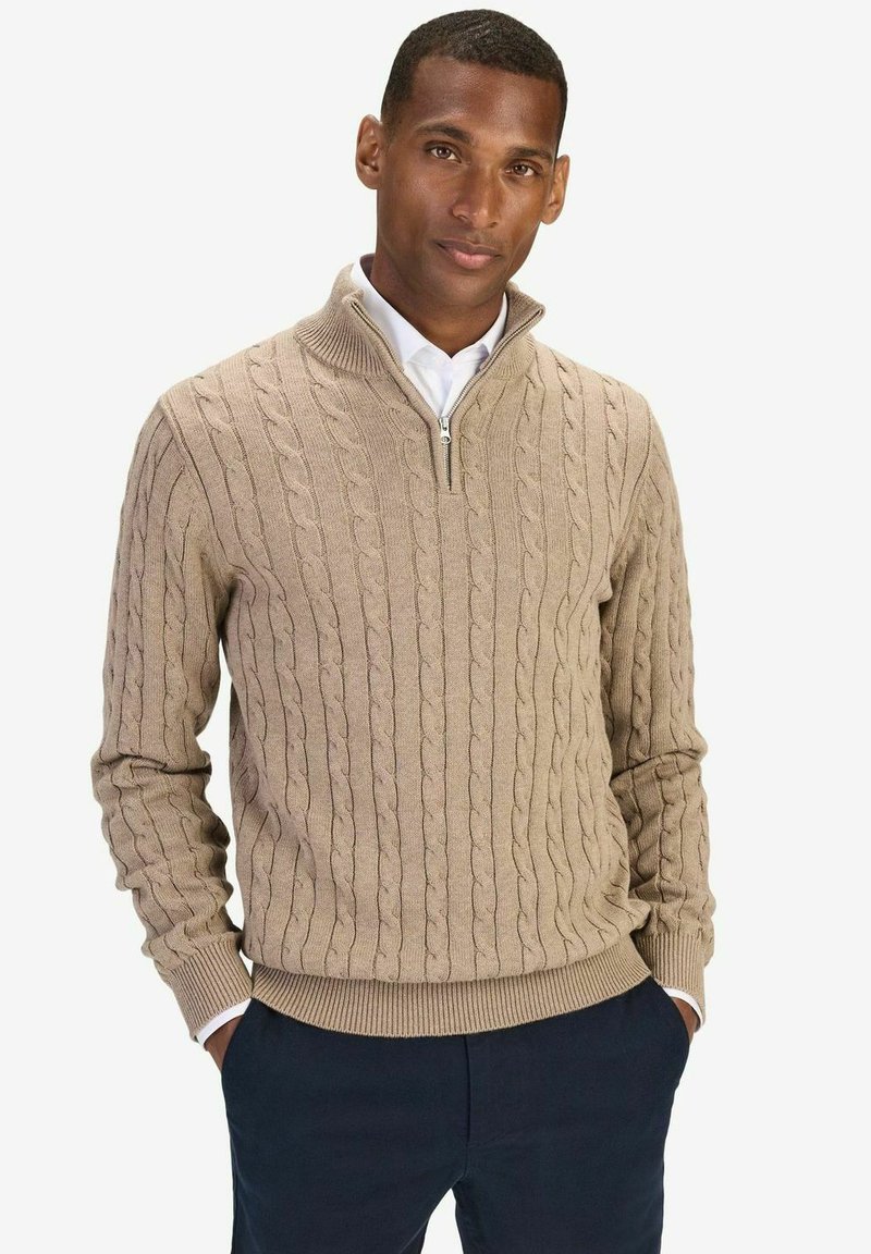 Beige kabelstickad tröja med halvzip-krage, med texturerade mönster och ribbade ärmslut, i kombination med marinblå byxor.