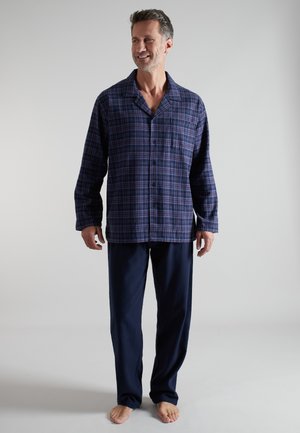 Man smiling, wearing blue plaid buttoned pajama top and solid navy pajama pants, standing barefoot against plain light background.
