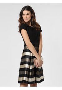 Schwarzes Oberteil mit rundem Ausschnitt, kombiniert mit einem knielangen Rock, der abwechselnd schwarze und metallisch goldene Streifen aufweist. Hält eine silberne Clutch.