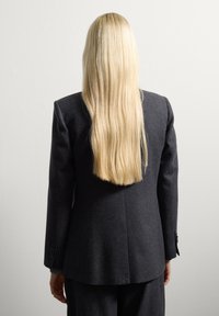 Chaqueta gris oscuro a medida con mangas largas, solapa con muesca y textura sutil, vista desde la parte de atrás con cabello rubio largo y liso visible.