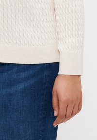 Kremfarget teksturert genser med kabelstrikkmønster og ribbestrikkede mansjetter, iført over blå denim, med en hånd hvilende ved siden av midjen.
