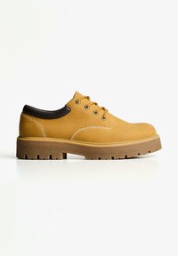 Pantofi bej cu șireturi, din suede, cu o guleră din piele închisă la culoare, având o talpă groasă maro și cusături albe pe margine.