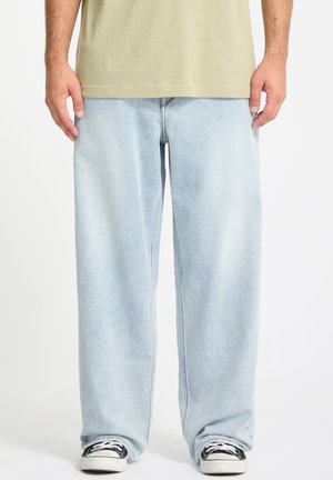 Personne portant un jean large bleu clair, un t-shirt beige, et des baskets noires et blanches à lacets, debout sur un fond blanc.