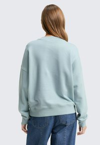 Világoskék kapucnis pulóver, amely laza szabású, bordás nyakkivágással és mandzsettákkal rendelkezik, valamint puha textúrájú. Közepesen koptatott farmernadrággal párosítva.