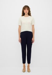 Kurzarmpullover aus cremefarbenem Strick mit strukturiertem Muster, kombiniert mit dunkelblauen, maßgeschneiderten Hosen und hautfarbenen Slippern.