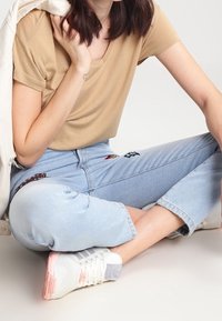 T-shirt beige a scollo a V abbinato a jeans chiari, leggermente corti, con toppe colorate. Sneakers bianche con suola rosa.
