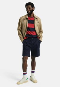 Khakifarbene Jacke über einem rot-blauen gestreiften T-Shirt, kombiniert mit marineblauen Shorts. Weiße Turnschuhe mit grünen Akzenten und gestreiften Socken. Lässige Kleidung.