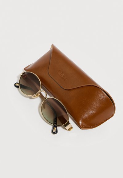 Στρογγυλά γυαλιά ηλίου με χρυσή κορνίζα και καφέ φακούς με διαβάθμιση, δίπλα σε καφέ δερμάτινη θήκη γυαλιών ηλίου Chloé σε λευκή επιφάνεια.