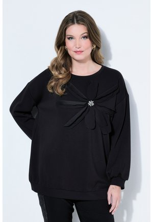 Kobieta o falowanych włosach ubrana w czarny top z długim rękawem, zdobiony dużym dekoracyjnym kwiatem z tkaniny z błyszczącym, ozdobnym środkiem.