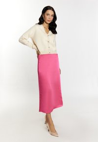 Cardigan crema con dettagli sui bottoni, abbinato a una gonna midi in satin rosa. Modella che indossa tacchi alti nudi, capelli acconciati in morbide onde.