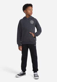 Felpa grigia con tasca frontale e logo, abbinata a pantaloni tuta neri e sneaker nere con suole bianche. Tessuto testurizzato, vestibilità rilassata.