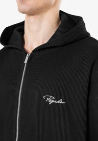 Black ribbed zip-up hoodie with a large, structured hood and a white embroidered logo on the chest. The fabric has a textured appearance.