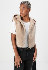 Bež prsluk bez rukava s kapuljačom, sa prednjim dijelom sličnim krznu i glatkom stražnjom stranom. Zatvaranje na patentni zatvarač i nosi se s bijelom majicom i tamnim trapericama.