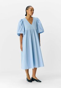 Robe bleu clair à décolleté en V avec des manches courtes à volants et une taille froncée, offrant une coupe ample s'étendant jusqu'au mi-mollet. Des ballerines noires complètent le look.