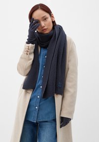 Écharpe et gants en tricot côtelé bleu marine portés avec un manteau beige sur une chemise en denim, arborant des accents de boutons sur la manche du manteau.
