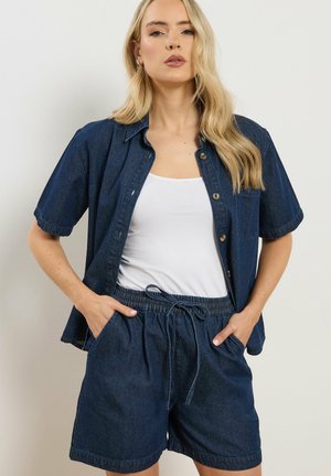 Frau trägt dunkelblaues Jeanshemd mit kurzen Ärmeln über weißem Tanktop, kombiniert mit passenden Jeans-Shorts mit Kordelzug, Hände in den Taschen.