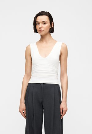 Jeune femme aux cheveux courts et foncés portant un haut blanc sans manches à col en V et un pantalon large foncé taille haute, debout devant un fond uni.