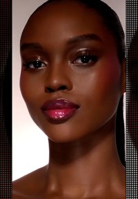 Close-up of a model's face, showcasing smooth dark skin, glossy pink lips, subtle blush, highlighted cheekbones, and defined eyes against a gradient background.