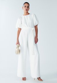Witte jumpsuit met wijde pijpen, met pofmouwen, een taille met ceintuur en een gladde textuur. Gecombineerd met gouden sandalen met bandjes en een metalen handtas.