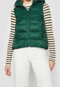 Padded zielona kamizelka z kapturem, z przodu na zamek błyskawiczny i regulowanymi paskami. Noszona na paski z długim rękawem oraz białe spodnie.