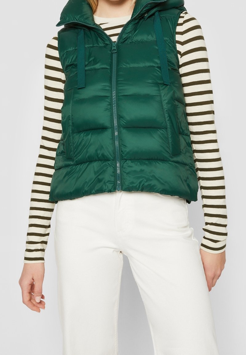 Padded zielona kamizelka z kapturem, z przodu na zamek błyskawiczny i regulowanymi paskami. Noszona na paski z długim rękawem oraz białe spodnie.