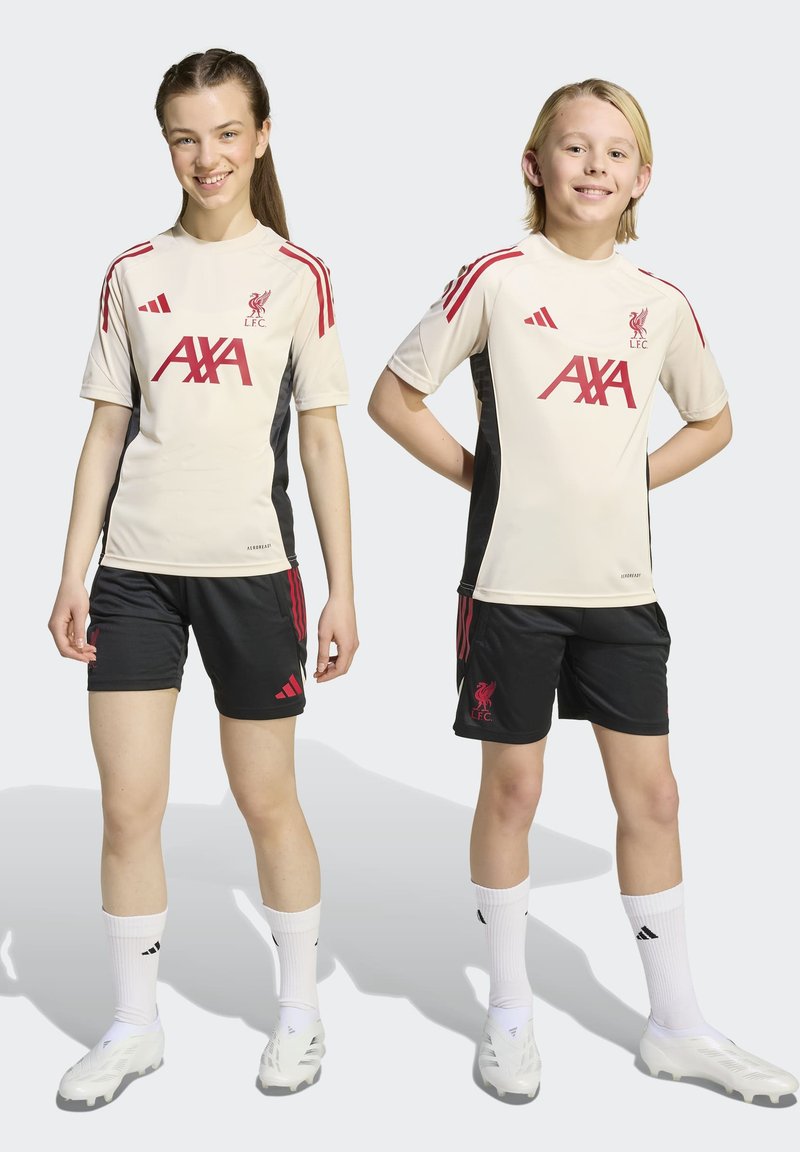 Zwei Kinder in beigen und schwarzen Liverpool FC Fußballtrikots mit weißen Fußballschuhen, die nebeneinander vor einem einfarbigen Hintergrund stehen.