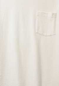 T-shirt de couleur crème claire en tissu texturé, avec une seule poche poitrine dotée d'un détail de bordure cousue.