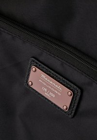 Sac en tissu noir avec une étiquette en cuivre portant le logo "Maserati Piquadro", numéroté 176/200, fermeture éclair sécurisée et surface texturée.