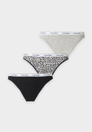 Femme debout pieds nus portant un bralette blanc Calvin Klein et un slip noir Calvin Klein sur un fond clair uni.