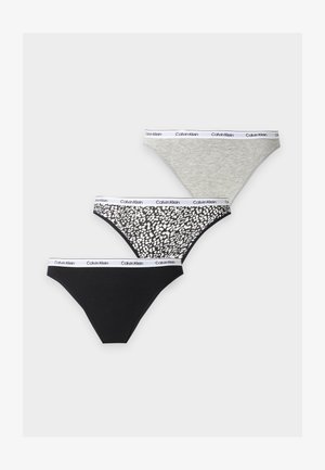 Kobieta stoi boso, ubrana w biały bralet Calvin Klein i czarne majtki Calvin Klein na jednolitym jasnym tle.