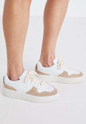 Person wearing white sneakers with beige suede accents and thick soles, standing on a light neutral surface.
