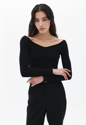 Maglietta nera a maniche lunghe con scollo a V ampio. Design aderente, texture liscia e stile minimalista. Abbinata a pantaloni neri.