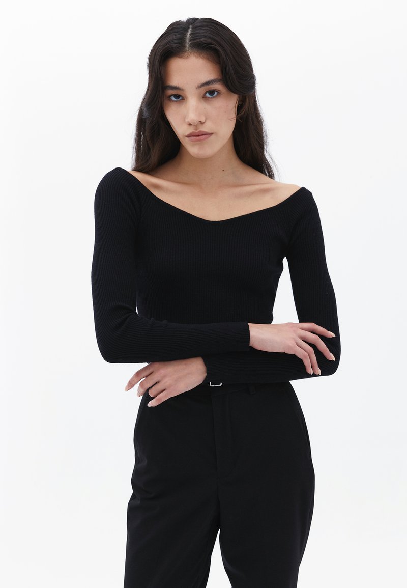 Schwarzes, geripptes Langarmoberteil mit weitem V-Ausschnitt. Figurumspielendes Design, glatte Textur und minimalistischer Stil. Kombiniert mit schwarzen Hosen.