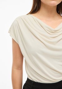 Kvinne iført en kremfarget drapert bluse stukket inn i svarte bukser, vist fra skuldrene til midjen mot en hvit bakgrunn.