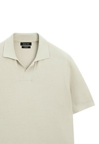 Ljust beige bomullspolo med strukturerad väv, korta ärmar och V-ringning. Logotyp från varumärket syns innanför kragen.